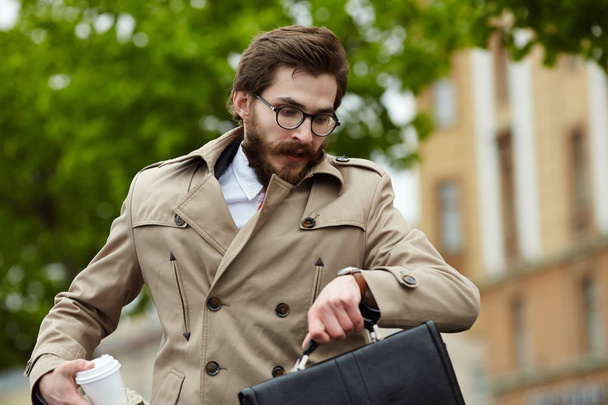 Hurrying businessman in elegant trench looking at watch in urban environment - Foto, Imagen