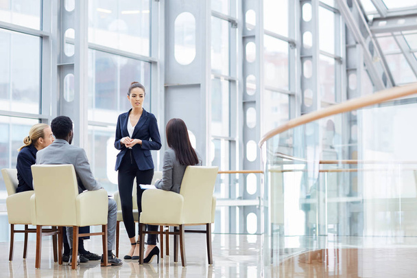 会議では、近代的なオフィスビルのガラスのホールでプレゼンテーションを与えて 1 つの女性の間に肘掛け椅子に座っている 4 つのビジネス人々 のグループ - 写真・画像