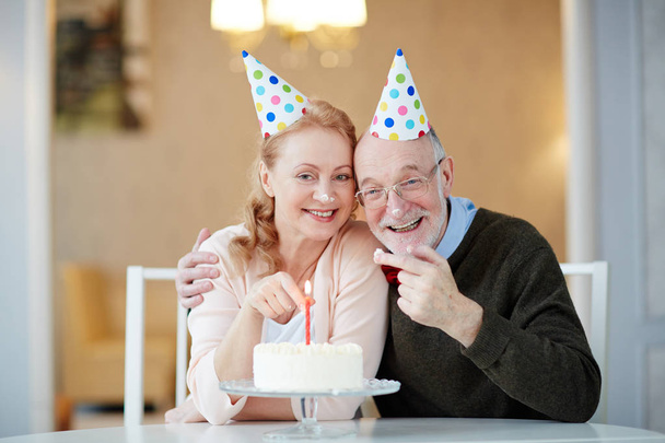 Cheerful husband and wife in birthday caps tasting cake - Fotoğraf, Görsel