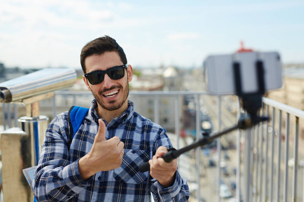 Retrato de jovem bonito mostrando polegares até a câmera tirando foto selfie enquanto estava em pé no telhado contra a vista panorâmica da cidade e binóculos operados por moedas no fundo
 - Foto, Imagem