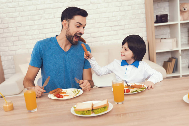 A man spends time with his son. The father of boy is engaged in raising child. Father feeds his son. The boy is having breakfast with his father. - Фото, изображение
