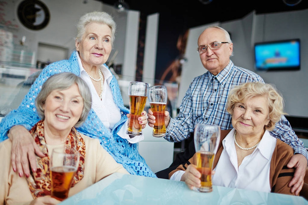 Čtyři starší přátelé v hospodě: oni sedí u stolu s pivní sklenice a pózuje pro fotografie, jeden z nich koukal zamyšleně - Fotografie, Obrázek