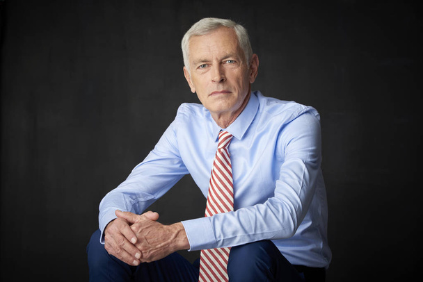 Confident senior man wearing shirt and tie and looking thoughtful while sitting before dark grey wall. - Photo, image