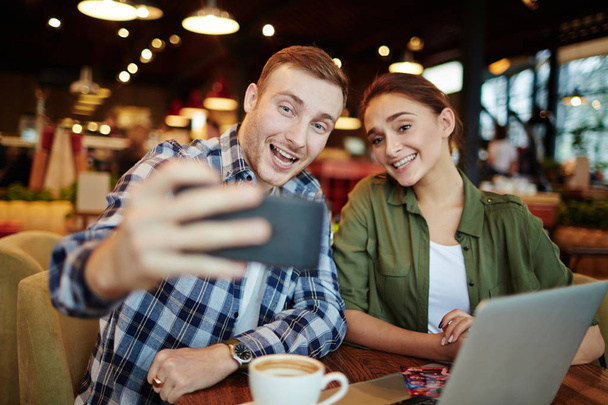 Pozostawiając w pamięci ciepły wieczór z przyjacielem: całkiem młoda kobieta i przystojny Brodaty mężczyzna biorąc selfie na smartphone, siedząc w przytulnej kawiarni - Zdjęcie, obraz