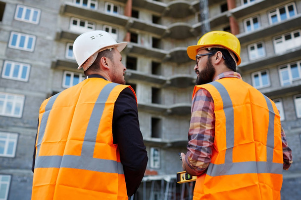 Ruggen van twee bouwers in uniform onder discussie over achtergrond van onvoltooide bouwwerk - Foto, afbeelding