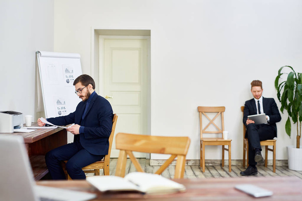 Deux hommes d'affaires travaillant dans un bureau d'affaires de la vieille école, un homme lisant la documentation au bureau, un autre utilisant une tablette assise sur une chaise
 - Photo, image