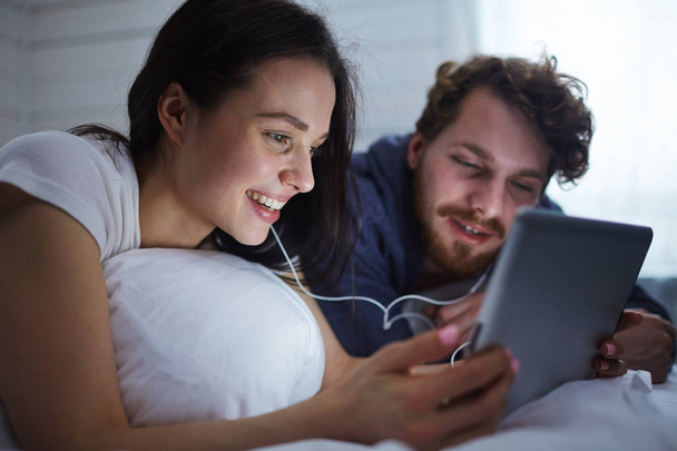 Σύγχρονο νεαρό ζευγάρι βλέποντας ταινία στο touchpad ξαπλωμένοι στο κρεβάτι - Φωτογραφία, εικόνα