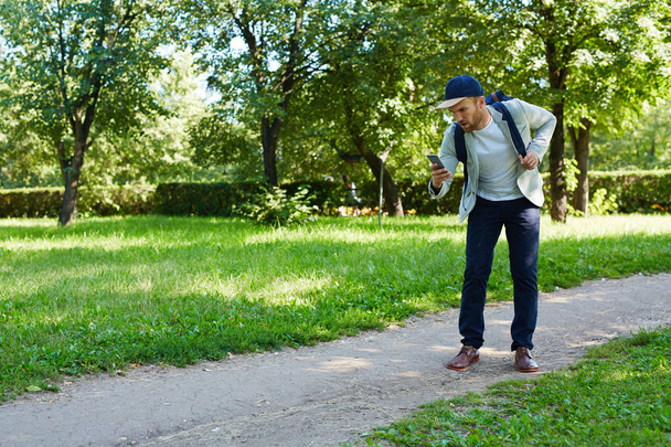 Ganzkörperporträt eines gutaussehenden jungen Mannes, der mit Rucksack in einem sonnigen öffentlichen Park spaziert und auf dem Smartphone im Internet surft - Foto, Bild