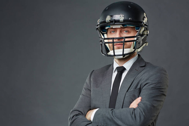 Fiducioso uomo d'affari in giacca e cravatta e casco da football americano in piedi con le braccia incrociate sullo sfondo grigio e guardando la fotocamera, copiare lo spazio a sinistra
 - Foto, immagini
