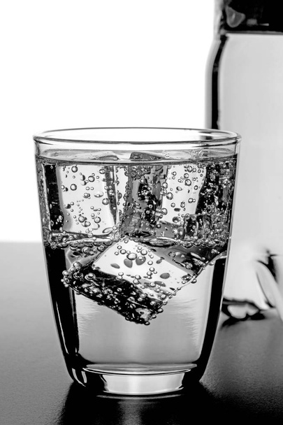 Een glas koud water met ijsblokjes en een fles in de achtergrond - Foto, afbeelding