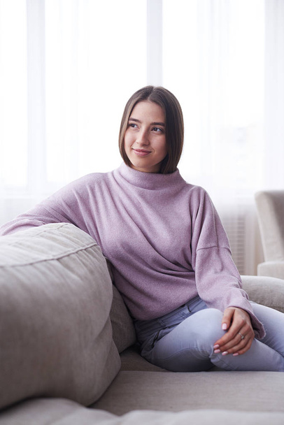 Κοπέλα φορώντας άνετο πουλόβερ που κάθεται στον καναπέ στο σπίτι, απολαμβάνοντας leis - Φωτογραφία, εικόνα