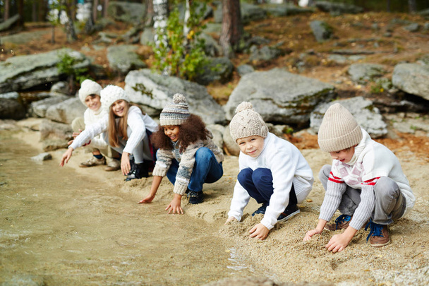 Skupina šťastných dětí hraní venku v přírodě: sedí na břehu jezera, které se dotýkají pískem a průzračnou vodou na teplý podzimní den - Fotografie, Obrázek