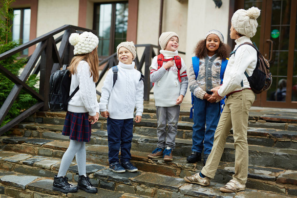 Skupina dětí chatování stojící na prahu školy, všichni nosí batohy a podobné pletené oblečení - Fotografie, Obrázek
