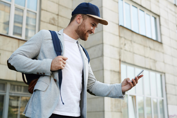 Profilbild eines fröhlichen jungen Mannes, der mit seinem Freund auf dem Smartphone SMS schreibt, während er vor einem mehrstöckigen Gebäude steht, Taille-up-Porträt - Foto, Bild