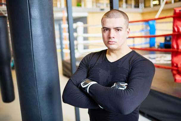 Retrato de homem muscular confiante posando com saco de perfuração e olhando para a câmera durante a prática de boxe no clube esportivo
 - Foto, Imagem