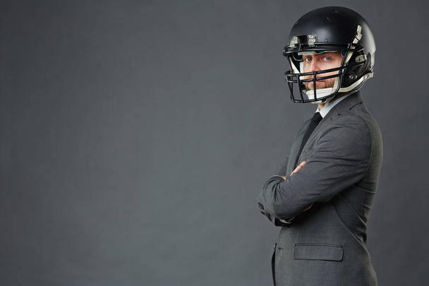 Side view of middle adult businessman wearing American football helmet standing cross-armed against grey background and looking at camera, copy space to left - Photo, Image