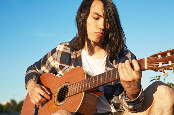 Portrét jistý muž v asijské kostkovaná košile hrál na kytaru zatímco sedí venku, bezmračné modré obloze na pozadí - Fotografie, Obrázek