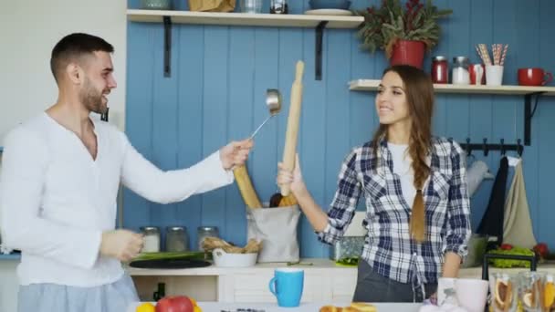 Happy couple having fun in the kitchen fencing with ladle and rolling-pin while cooking breakfast at home - Кадри, відео