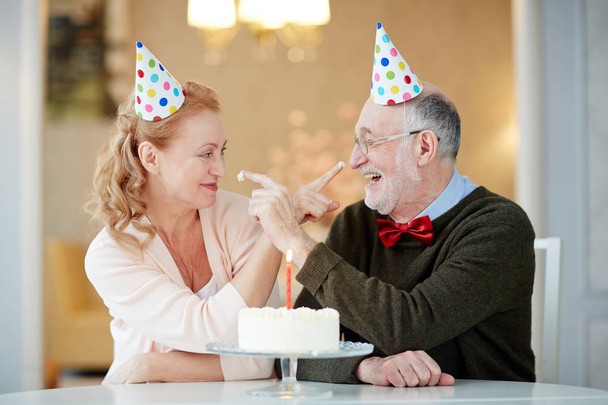 楽しい遊び心のあるシニア カップルの肖像画の誕生日ケーキとテーブルで一緒に座っているとクリームとお互いを汚れしようとしてのパーティの帽子を身に着けているを祝う  - 写真・画像