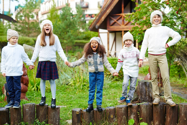 fröhliche Kinder unterschiedlichen Alters, die Spaß daran haben, draußen in der grünen Landschaft zu spielen, in Reihen auf Holzpfählen stehend und Hände in ähnlich weißer Strickkleidung haltend - Foto, Bild