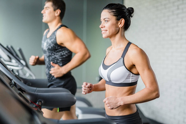 Uygun adamın yanında spor salonunda treadmill egzersiz yaparken gülümseyen güzel sportif esmer kadın portresi - Fotoğraf, Görsel