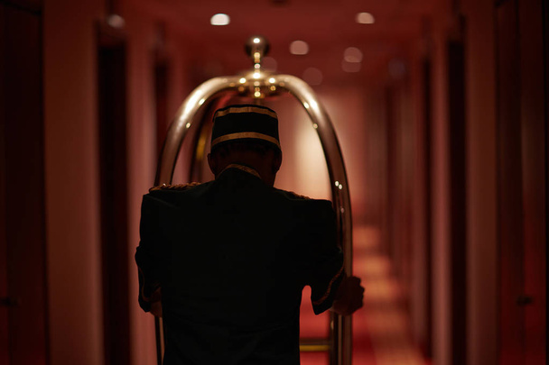 Vista trasera del sirviente moviéndose a lo largo del pasillo del hotel con carrito de equipaje
 - Foto, Imagen