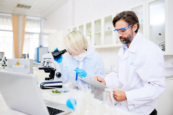 Вчений у білому костюмі робить нотатки, а його колега вивчає хімічну речовину в мікроскопі
 - Фото, зображення
