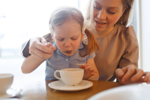 Adorable child sipping hot drink from spoon with her mother  - Foto, Imagen