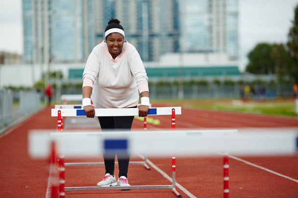 Счастливая женщина в спортивной одежде во время тренировок на стадионе наклоняется вперед на препятствиях
 - Фото, изображение