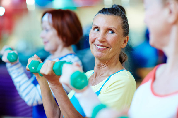 Smiley mulher madura com halteres e seus amigos se exercitando no ginásio
 - Foto, Imagem