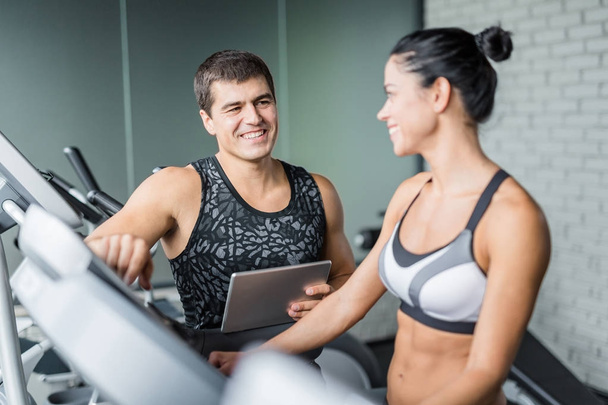 Modern bir spor salonu egzersiz için uygun kadın kullanım eliptik makinesi gülümsüyor izlerken kas kişisel eğitmen portresi - Fotoğraf, Görsel