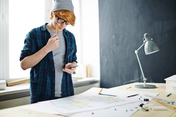 Porträt eines jungen rothaarigen Designers mit Brille und Mütze bei der Arbeit im Büro: am Schreibtisch mit Grundrissen stehen, denken und Smartphone benutzen - Foto, Bild