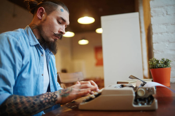 Side view portrait of creative tattooed man using old-school vintage typewriter on wooden table in cafe, typing story looking for inspiration - Photo, Image