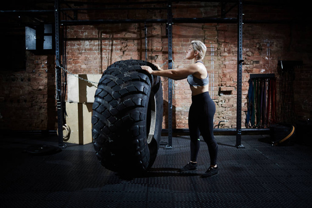 Vista del profilo di una donna piuttosto sportiva che ribalta un pneumatico enorme durante una sessione di allenamento intensivo in palestra
 - Foto, immagini