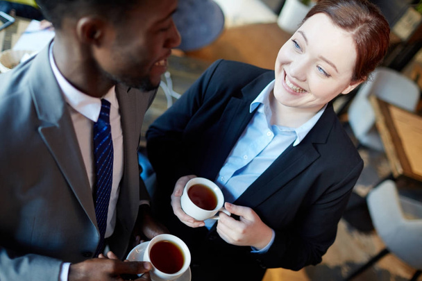 Angolo alto di due uomini d'affari che interagiscono durante la pausa caffè: Giovane donna che parla con un collega afro-americano sorridente e con le tazze in mano con il tè
  - Foto, immagini