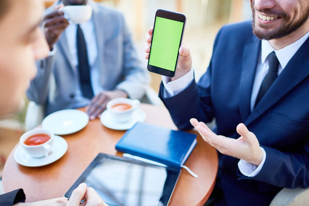 Closeup portret van lachende zakenman presenteert smartphone met groene leeg scherm aan collega's tijdens vergadering hen mobiele toepassing delen uit te nodigen - Foto, afbeelding