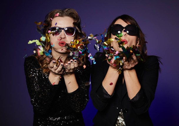 Trendy young women in sunglasses blowing colorful confetti while standing against dark background, waist-up group portrait - Fotó, kép
