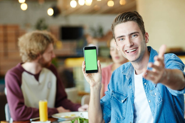 Μέση-κατακόρυφα χαρούμενα νεαρού άνδρα που δείχνει στο camera κρατώντας στο χέρι, στο σύγχρονο smartphone τους φίλους του να κουβεντιάζουν ζωηρά με το άλλο και απολαμβάνοντας το μεσημεριανό γεύμα στο ζεστό καφέ - Φωτογραφία, εικόνα