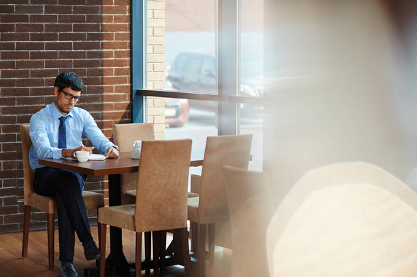 Koncentrovaná vousatý podnikatele nezbytné poznámky při posezení v útulné kavárně s panoramatickými okny, celovečerní portrét - Fotografie, Obrázek