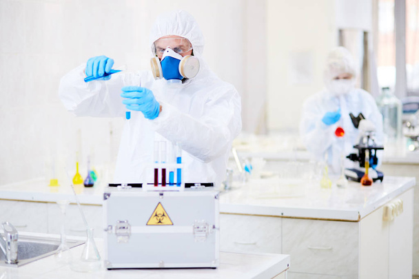 Mies tutkija yllään hazmat puku lisäämällä sininen ratkaisu koeputkeen suorittaessaan kokeilun nykyaikaisessa laboratoriossa
 - Valokuva, kuva