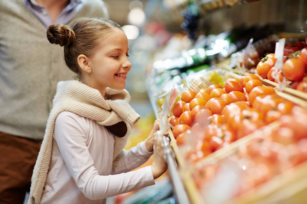 Retrato de vista lateral de la linda niña sonriendo inclinada sobre el mostrador de verduras con tomates maduros frescos y otras verduras
 - Foto, imagen