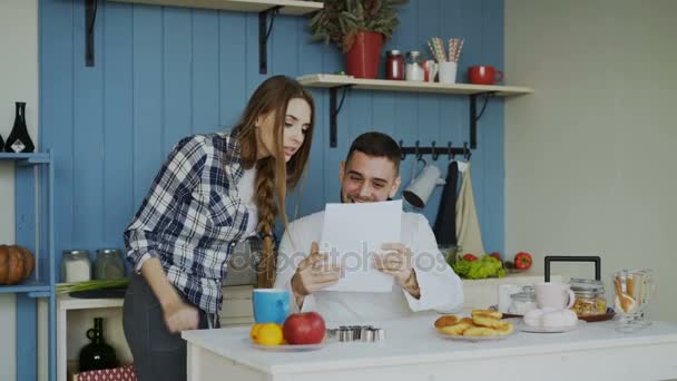 Attractive happy couple recieve good news unfolding letter in the kitchen while have breakfast at home - Video