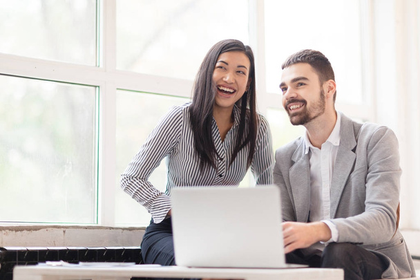 Pretty Asian office worker and her handsome colleague distracted from working on laptop and looking away with toothy smiles, waist-up portrait - Foto, Bild