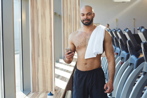 Mięśni shirtless sportowca z gadżet patrząc na kamery w siłowni - Zdjęcie, obraz