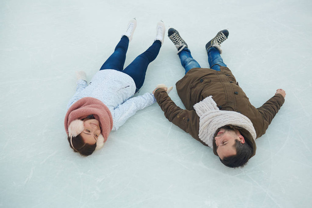 Szczęśliwi zakochani w winterwear leżącego na lodowisko i trzymając się za ręce - Zdjęcie, obraz