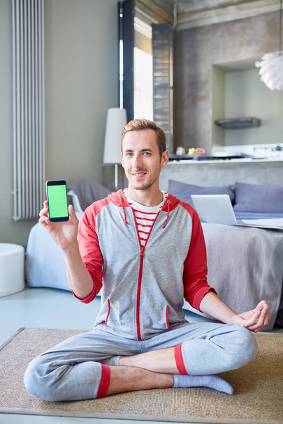 Νέος άνθρωπος με smartphone δείχνει περίεργο promo ενώ κάθεται σε στάση του λωτού, στο πάτωμα του υπνοδωματίου - Φωτογραφία, εικόνα