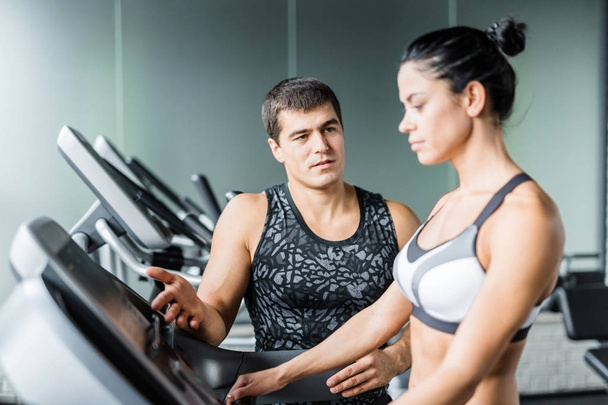 Portrait of muscular personal instructor explaining to fit woman how to set up elliptical machine for exercise in modern gym - Photo, Image