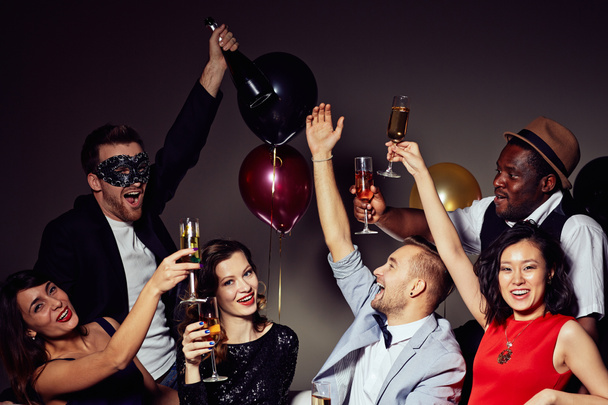 Verjaardagsfeestje thuis hebben: vrolijke vrienden op zoek naar camera met toothy glimlacht terwijl rammelende champagne samen, kleurrijke ballonnen op achtergrond fluiten - Foto, afbeelding