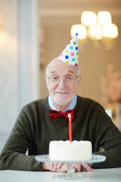 Alter Mann mit Geburtstagsmütze sitzt am Tisch vor Kuchen mit brennender Kerze - Foto, Bild