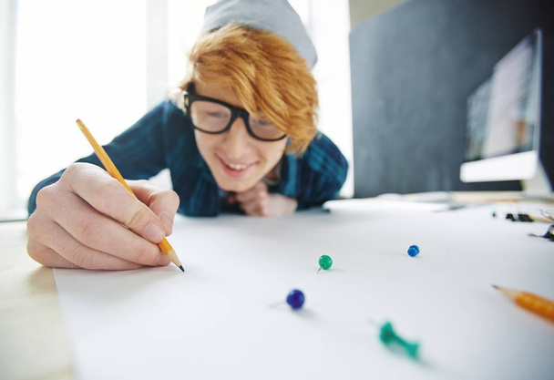 Retrato de un joven creativo pelirrojo con gorro y gafas dibujando con lápiz apoyado cerca del escritorio y sonriendo alegremente
 - Foto, Imagen